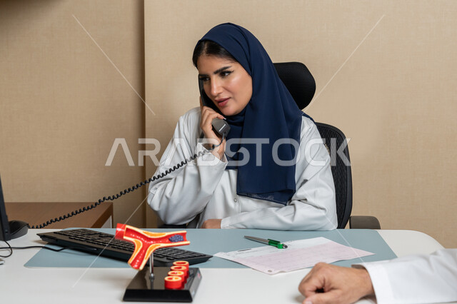 Providing medical services to patients with all efficiency, women's professions and jobs, close-up image of an Emirati Gulf Arab female doctor sitting behind her desk making a phone call to inquire about something, the concept of medicine and health care