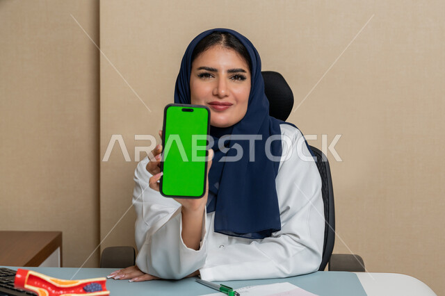 التواصل مع المرضى عبر الهاتف المحمول، مهن ووظائف نسائية، طبيبة عربية خليجية اماراتية محجبة ترتدي المعطف الطبي تجلس خلف الطاولة تقوم بعرض شاشة خضراء على هاتفها، العمل في القطاع الاماراتي الصحي، استخدام القنيات الحديثة المتطورة في المجال الطبي