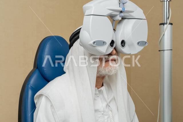 تشخيص وتحديد المشاكل البصرية، صورة مقربة لرجل مسن عربي اماراتي خليجي يرتدي الغترة امام جهاز الفحص البصري للتأكد من صحة النظر، تقييم مدى الرؤية والكشف عن الامراض العينية، مفهوم الخدمات والرعاية الصحية في المستشفيات والمراكز، استخدام الأجهزة الحديثة المتطورة