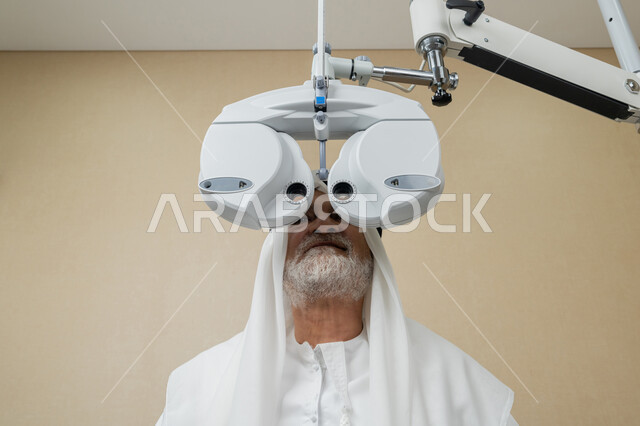 تشخيص وتحديد المشاكل البصرية، صورة مقربة لرجل مسن عربي اماراتي خليجي يرتدي الغترة امام جهاز الفحص البصري للتأكد من صحة النظر، تقييم مدى الرؤية والكشف عن الامراض العينية، مفهوم الخدمات والرعاية الصحية في المستشفيات والمراكز، استخدام الأجهزة الحديثة المتطورة