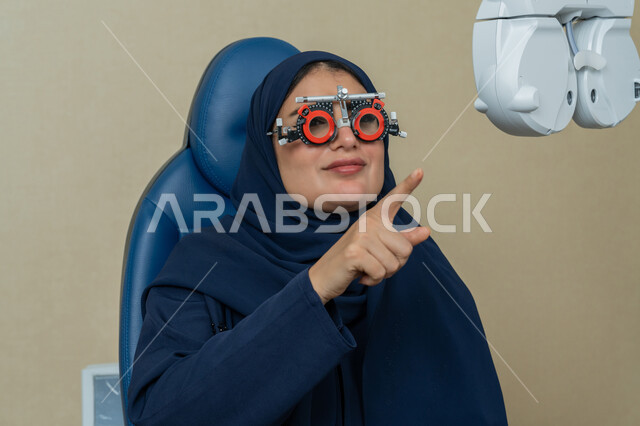 الاشارة الى شيء ما باصصبع السبابة، استخدام الأجهزة الحديثة المتطورة، تشخيص وتحديد المشاكل البصرية، صورة مقربة لامراة عربية خليجية اماراتية ترتدي العباءة تجلس أمام جهاز فحص العين للتأكد من صحة البصر، تقييم مدى الرؤية والكشف عن الامراض العينية، مفهوم الخدمات والرعاية الصحية