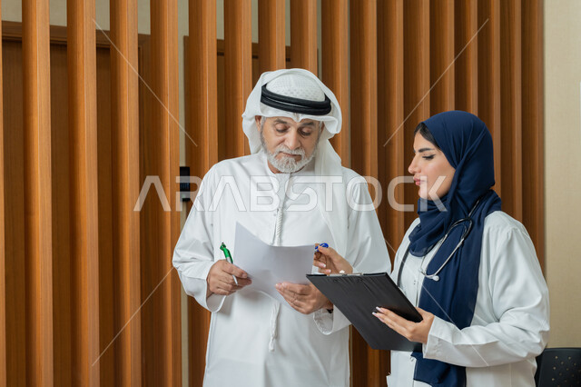 تقديم الخدمات والاستشارات الطبية للمراجعين، مفهوم الطب والرعاية الصحية، طبيبة عربية خليجية اماراتية محجبة ترتدي المعطف الابيض تقوم بتدوين المعلومات العامة عن حالة مريض مسن اماراتي، مهن ووظائف نسائية، دراسة و مراجعة حالات المرضى