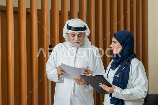 مفهوم الطب والرعاية الصحية، تقديم الخدمات والاستشارات الطبية للمراجعين، طبيبة عربية خليجية اماراتية محجبة ترتدي المعطف الابيض تقوم بتدوين المعلومات العامة عن حالة مريض مسن اماراتي، مهن ووظائف نسائية، دراسة و مراجعة حالات المرضى