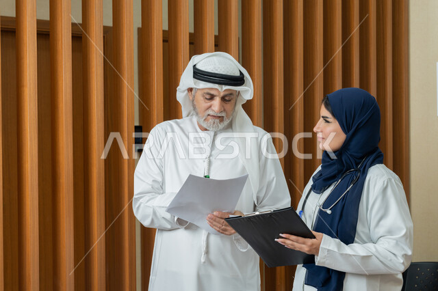 Providing medical services and consultations to patients, the concept of medicine and health care, a veiled Gulf Arab Emirati female doctor wearing a white coat recording general information about the condition of an elderly Emirati patient, women's professions and jobs, studying and reviewing patients' cases