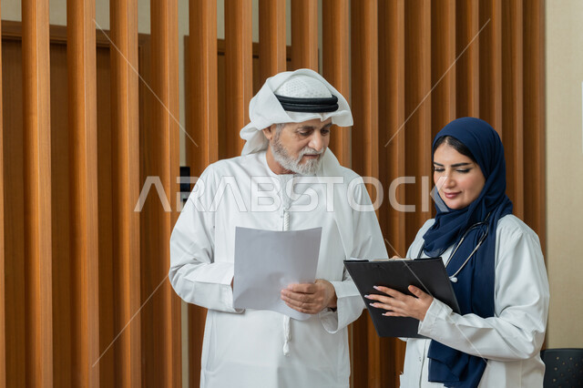 تقديم الخدمات والاستشارات الطبية للمراجعين، مفهوم الطب والرعاية الصحية، طبيبة عربية خليجية اماراتية محجبة ترتدي المعطف الابيض تقوم بتدوين المعلومات العامة عن حالة مريض مسن اماراتي، مهن ووظائف نسائية، دراسة و مراجعة حالات المرضى