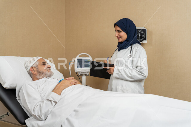 The concept of medicine and health care, providing medical services and consultations to patients, an Arab Gulf Emirati female doctor wearing a hijab and a white coat is recording information about an elderly Arab Emirati patient lying on the bed, women's professions and jobs, studying and reviewing patients' cases