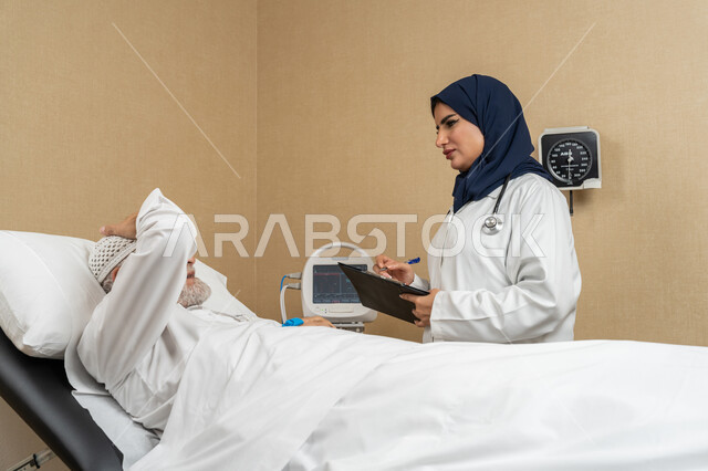 تقديم الخدمات والاستشارات الطبية للمراجعين، مفهوم الطب والرعاية الصحية، طبيبة عربية خليجية اماراتية ترتدي الحجاب والمعطف الابيض تقوم بتدوين معلومات مريض مسن عربي اماراتي مستلقي على السرير، مهن ووظائف نسائية، دراسة و مراجعة حالات المرضى