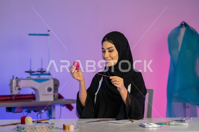 أهمية المرأة في مجال الخياطة، موديلات جديدة وعصرية، بورتريه مقرب لامرأة عربية خليجية امارتية ترتدي العباءة تحمل بيدها بكرة من الخيطان، ورشة عمل تصميم ازياء، مهن وأعمال فنية نسائية، شابة سعودية تقوم تفصيل الملابس الراقية، مفهوم عالم الأزياء والموضة، خلفية ملونة