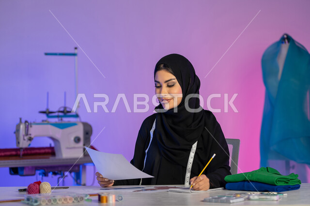 أعمال ومهن فنية يدوية, مهارة تفصيل الملابس النسائية، اختراع وابتكار موديلات جديدة وعصرية، بورتريه مقرب لمصممة أزياء عربية خليجية إماراتية ترتدي العباءة تستخدم معدات الرسم والخياطة, شابة سعودية تعمل باندماج, خلفية ملونة