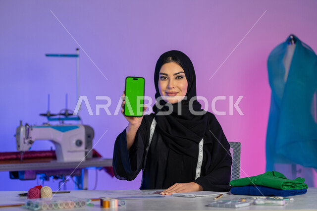 فنون تفصيل الملابس النسائية, استخدام الأجهزة والتقنيات الحديثة, شابة سعودية تعمل عبر الأونلاين, عرض شاشة خضراء فارغة على الهاتف المحمول, دمج العمل مع التقنية والتكنولوجيا, بورتريه مقرب لمصممة عربية خليجية إماراتية ترتدي العباءة تنظر إلى الكاميرا, خلفية ملونة