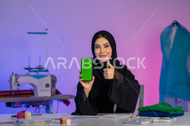 تعابير الاعجاب والامتياز والجودة, عرض شاشة خضراء فارغة على الهاتف المحمول, دمج العمل مع التقنية والتكنولوجيا, بورتريه مقرب لمصممة عربية خليجية إماراتية ترتدي العباءة تنظر إلى الكاميرا, فنون تفصيل الملابس النسائية, شابة سعودية تعمل عبر الأونلاين, خلفية ملونة