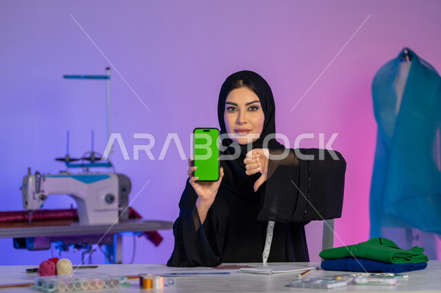 رفع الاصبع الابهام للأسفل بعلامة الرفض وعدم الاعجاب, عرض شاشة خضراء فارغة على الهاتف المحمول, دمج العمل مع التقنية والتكنولوجيا, بورتريه مقرب لمصممة عربية خليجية إماراتية ترتدي العباءة تنظر إلى الكاميرا، شابة سعودية تقوم بفصيل الملابس النسائية, خلفية ملونة
