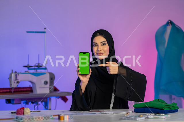 عرض شاشة خضراء فارغة على الهاتف المحمول, دمج العمل مع التقنية والتكنولوجيا, بورتريه مقرب لمصممة عربية خليجية إماراتية ترتدي العباءة تنظر إلى الكاميرا, فنون تفصيل الملابس النسائية, استخدام الأجهزة والتقنيات الحديثة, شابة سعودية تعمل عبر الأونلاين, خلفية ملونة