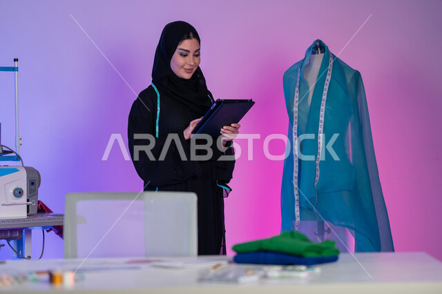معدات الخياطة ورسم الأزياء, دمج العمل مع التقنية والتكنولوجيا, بورتريه مقرب لمصممة أزياء عربية خليجية إماراتية تقف بجانب المانيكان وتعمل على جهاز التابليت في ورشة الخياطة, استخدام الأجهزة والتقنيات الحديثة والمتطورة, شابة سعودية تصمم موديلات جديدة, خلفية ملونة