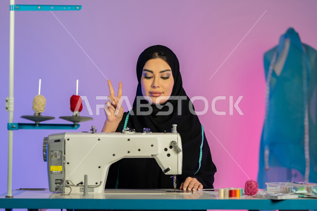 رفع الأصابع بعلامة التميز والجودة والرضا, تفصيل ملابس راقية, بورتريه مقرب لمصممة أزياء عربية خليجية اماراتية ترتدي العباءة تعمل على الماكينة, سيدة سعودية تجلس بإيماءات الثقة والاندماج, فنون وأعمال ومهن يدوية, الخياطة وتصميم الأزياء, خلفية ملونة