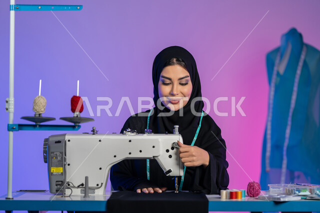 تنمية المهارات النسائية, ابتكار موديلات عصرية حديثة, تفصيل ملابس راقية, بورتريه مقرب لمصممة أزياء عربية خليجية اماراتية ترتدي العباءة تعمل على الماكينة, سيدة سعودية تجلس بإيماءات الثقة والاندماج, فنون وأعمال ومهن يدوية, فن الخياطة وتصميم الأزياء, خلفية ملونة