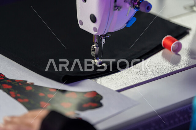 Professions and artistic works, arts and skills of tailoring women's clothing, concept of the world of fashion and style, close-up image of the hands of an Arab Gulf Emirati fashion designer sewing clothes on a machine, a young Saudi woman working in a fashion design workshop, sewing and knitting tools