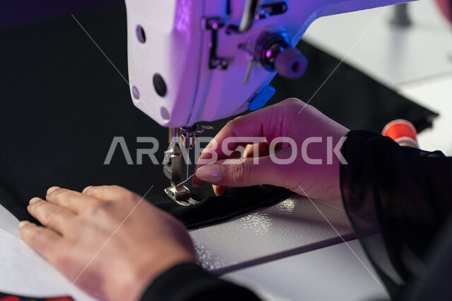أدوات الخياطة والحياكة، مهن وأعمال فنية، فنون ومهارات تفصيل الملابس النسائية، مفهوم عالم الأزياء والموضة, صورة مقربة لأيدي مصممة أزياء عربية خليجية إماراتية تقوم بخياطة الثياب على الماكينة, شابة سعودية تعمل في ورشة تصميم ازياء