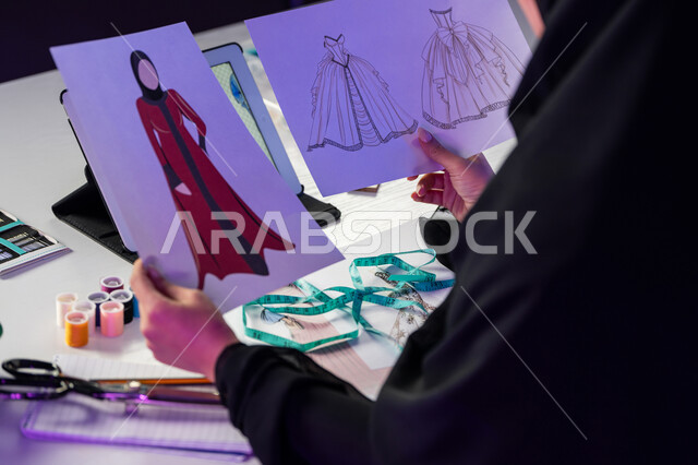 فن تصميم العباءات والفساتين العصرية, صورة مقربة من الجانب لمصممة أزياء عربية خليجية إماراتية ترتدي العباءة تحمل بيدها الرسومات, شابة سعودية تعمل في ورشة الخياطة, استخدام أدوات ومعدات تفصيل الملابس, مهارات وفنون نسائية يدوية