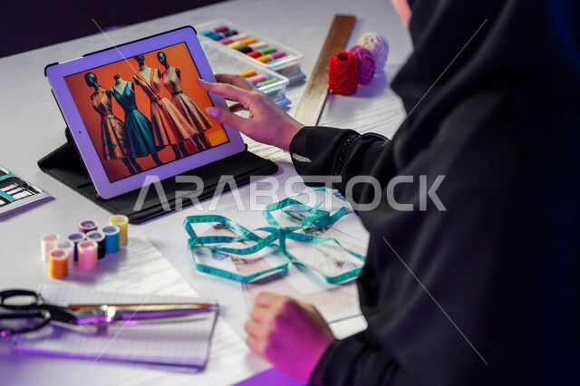 استخدام الأجهزة والتقنيات الحديثة, شابة سعودية تعمل عبر الأونلاين, أدوات ومعدات الخياطة والرسم, صورة مقربة من الجانب لمصممة أزياء عربية خليجية إماراتية ترتدي العباءة تقوم بتصفح التطبيقات عبر جهاز التابليت, فنون ومهارات تفصيل الملابس النسائية