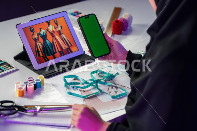 دمج العمل مع التقنية والتكنولوجيا, صورة مقربة من الجانب لمصممة أزياء عربية خليجية إماراتية ترتدي العباءة تقوم بعرض شاشة خضراء كروما على الهاتف المحمول, فنون ومهارات تفصيل الملابس النسائية, استخدام الأجهزة والتقنيات الحديثة, شابة سعودية تعمل عبر الأونلاين