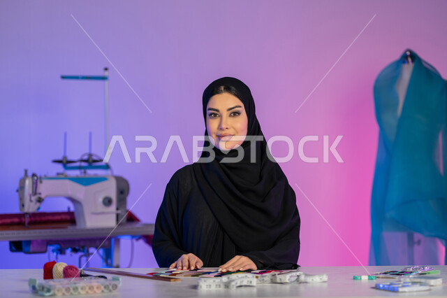 مهارة تفصيل الملابس النسائية، اختراع وابتكار موديلات جديدة وعصرية، بورتريه مقرب لمصممة أزياء عربية خليجية إماراتية ترتدي العباءة تستخدم معدات الرسم والخياطة, شابة سعودية تنظر إلى الكاميرا بثقة, مهن وأعمال فنية يدوية, خلفية ملونة