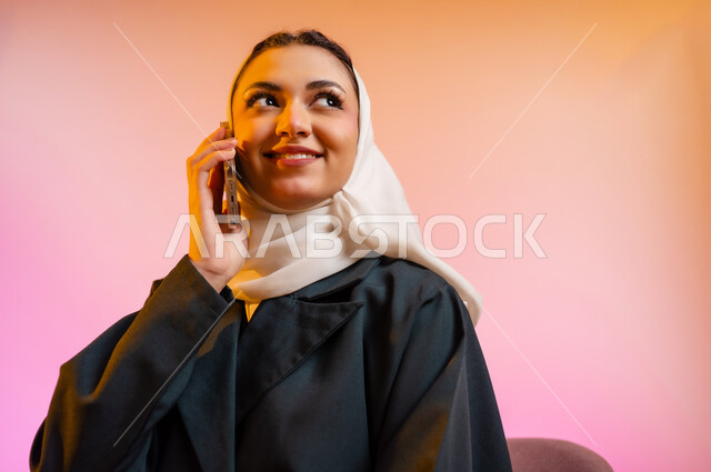 قضاء اوقات ممتعة بتبادل الاخبار والمحادثات عن بعد، بورتريه لسيده عربية خليجية عمانيه  محجبة  تقوم باجراء احاديث ودردشه مع الاهل والاصدقاء باستخدام الهاتف المحمول، ايماءات وجه تدل على السعادة و السرور، خلفيه ملونه