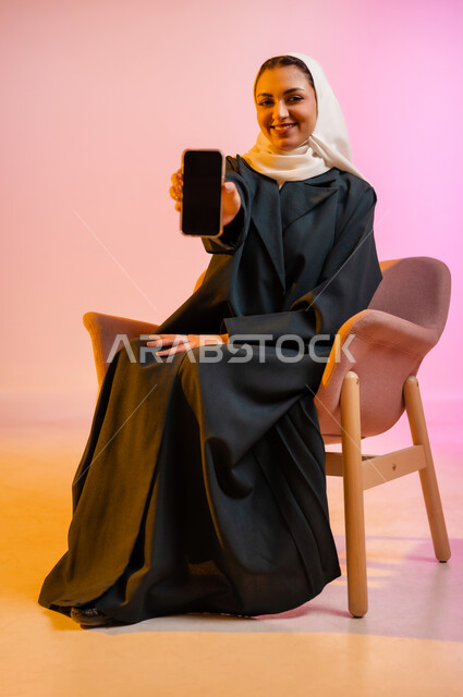 قضاء اوقات ممتعة بتبادل الاخبار والمحادثات عن بعد، بورتريه لسيده عربية خليجية عمانيه  محجبة  تمسك بيدها الهاتف المحمول وتشير به بشاشه سوداء فارغه، ايماءات وجه تدل على السعادة و السرور، خلفيه ملونه