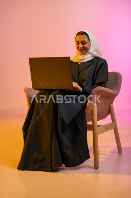 العمل لتحقيق الاهداف والاستراتيجيات، مهن ووظائف ادارية مكتبية, استخدام الأجهزة التكنولوجية الحديثة والمتطورة، إدارة شؤون الأعمال وتنظيمها، بورتريه لسيدة عربية خليجية عمانيه مبتسمه ترتدي الحجاب والعباءه تجلس عل كرسي مريح وتقوم بالعمل على جهاز الاب توب المحمول، خلفيه ملونه