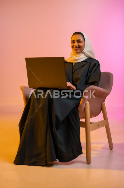 العمل لتحقيق الاهداف والاستراتيجيات، مهن ووظائف ادارية مكتبية, استخدام الأجهزة التكنولوجية الحديثة والمتطورة، إدارة شؤون الأعمال وتنظيمها، بورتريه لسيدة عربية خليجية عمانيه مبتسمه ترتدي الحجاب والعباءه تجلس عل كرسي مريح وتقوم بالعمل على جهاز الاب توب المحمول، خلفيه ملونه