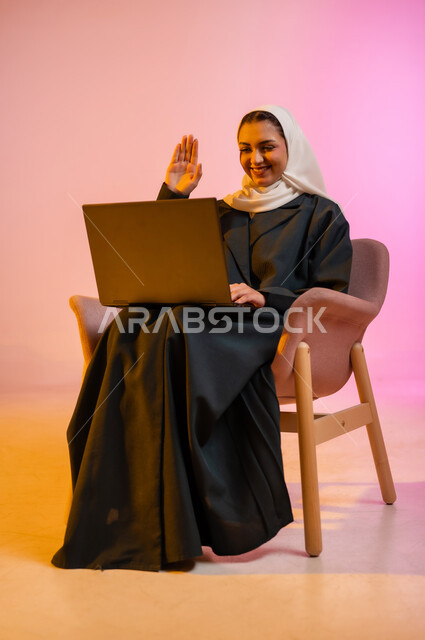 Working to achieve goals and strategies, office administrative professions and jobs, using modern and advanced technological devices, managing and organizing business affairs, portrait of a smiling Gulf Arab Omani woman wearing a hijab and abaya sitting on a comfortable chair and working on a portable laptop, colorful background