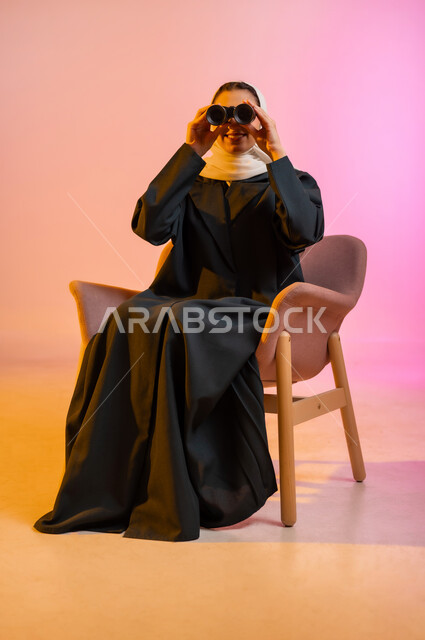 تقريب الاشياء البعيده بواسطه المنظار، بورتريه لسيدة عربية خليجية عمانيه ترتدي العباءه والحجاب تجلس على كرسي مريح وتمسك بيدها ناظور رؤية لمشاهدة شيئا ما بإيماءات و تعابير وجه و يدين تدلان على السعادة، الرؤية المستقبلية و التخطيط لرؤيه هدف ما، خلفيه ملونه