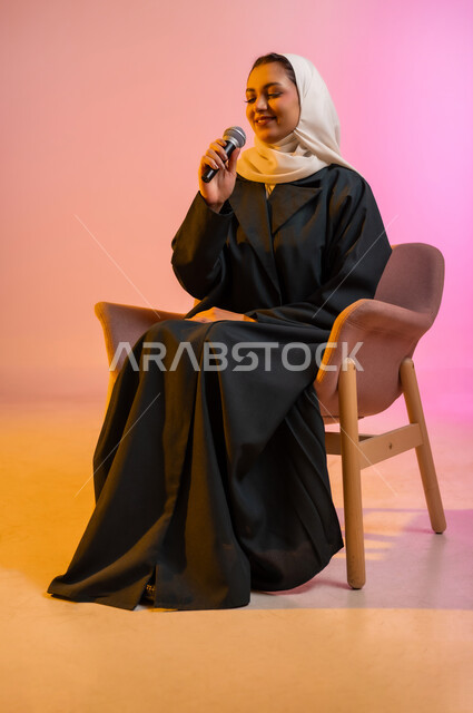 التدريب الصوتي وتقديم البرامج الاذاعيه، فن إلقاء الخطابات والندوات، بورتريه لشابة عربية خليجية عمانيه تجلس على كرسي مريح وتمسك بيدها الميكروفون، ايماءات وتعابير وجه و يدين تعبر عن الغناء والطرب، خلفية ملونه