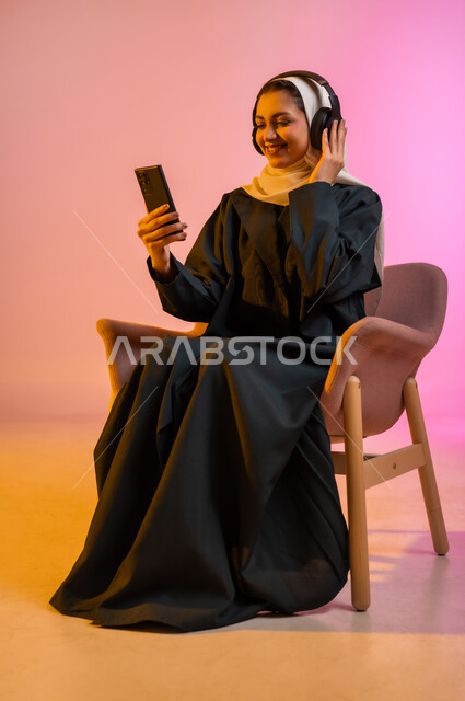 قضاء اوقات ممتعة بتبادل الاخبار والمحادثات عن بعد، بورتريه لسيده عربية خليجية عمانيه  محجبة  تقوم باجراء احاديث ودردشه مع الاهل والاصدقاء باستخدام الهاتف المحمول، ايماءات وجه تدل على السعادة و السرور، خلفيه ملونه