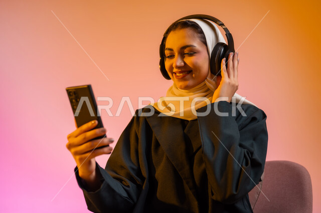 قضاء اوقات ممتعة بتبادل الاخبار والمحادثات عن بعد، بورتريه لسيده عربية خليجية عمانيه  محجبة  تقوم باجراء احاديث ودردشه مع الاهل والاصدقاء باستخدام الهاتف المحمول، ايماءات وجه تدل على السعادة و السرور، خلفيه ملونه