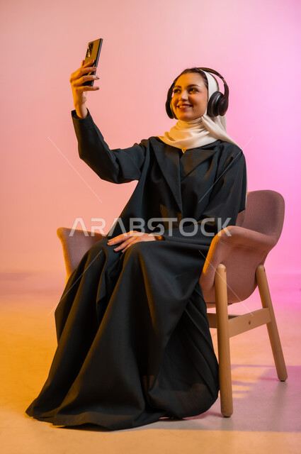 قضاء اوقات ممتعة بتبادل الاخبار والمحادثات عن بعد، بورتريه لسيده عربية خليجية عمانيه  محجبة  تقوم باجراء احاديث ودردشه مع الاهل والاصدقاء باستخدام الهاتف المحمول، ايماءات وجه تدل على السعادة و السرور، خلفيه ملونه