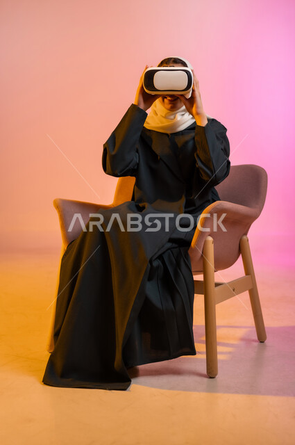 Technical and technological development, enjoying watching virtual reality movies and simulating the imaginary world, spending free time with recreational activities, portrait of a veiled Gulf Arab Omani woman wearing a colorful abaya sitting on a comfortable chair and looking through virtual reality glasses (3D VR), colorful background