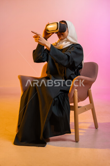التطور التقني والتكنولوجي, الاستمتاع بمشاهدة أفلام الواقع الافتراضي ومحاكاة العالم الخيالي, قضاء أوقات الفراغ بالأنشطة الترفيهية, بورتريه لسيده عربية خليجية عمانيه محجبة ترتدي العباءه الملونه تجلس على كرسي مريح وتنظر من خلال نظارة الواقع الافتراضي (في آر ثلاثية الأبعاد), خلفية ملونة
