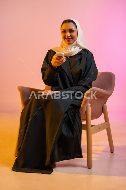 Remote control using modern electronic devices, a portrait of an Omani Gulf Arab woman wearing a colorful abaya and hijab, sitting on a comfortable chair, holding a remote control in her hand and pointing to a place, colorful background.