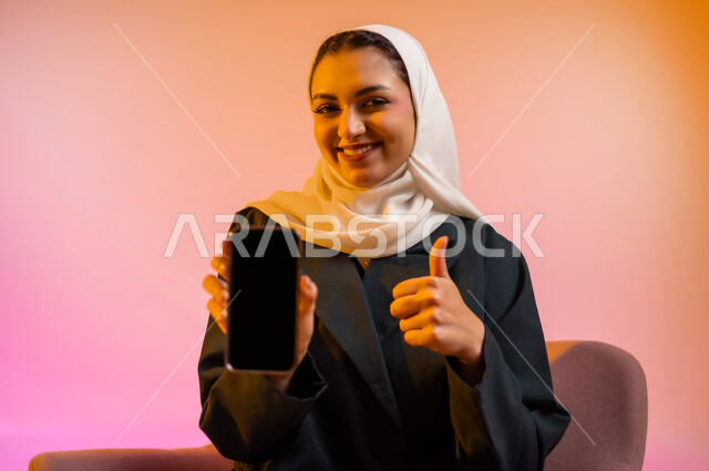 قضاء اوقات ممتعة بتبادل الاخبار والمحادثات عن بعد، بورتريه لسيده عربية خليجية عمانيه  محجبة  تمسك بيدها الهاتف المحمول وتشير به بشاشه سوداء فارغه، ايماءات وجه تدل على السعادة و السرور، خلفيه ملونه