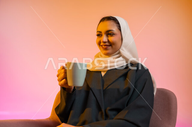 الإستمتاع بالمشروبات الدافئة واحتساء المشروبات الساخنه, بورتريه  لسيدة عربية خليجية عمانيه محجبة ترتدي العباءه الملونه تجلس على كرسي مريح وتقوم بشرب الشاي بواسطه كوب من السيراميك بابتسامه وطمأنينه، خلفيه ملونه