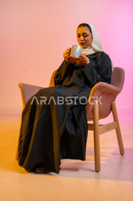 الإستمتاع بالمشروبات الدافئة واحتساء المشروبات الساخنه, بورتريه  لسيدة عربية خليجية عمانيه محجبة ترتدي العباءه الملونه تجلس على كرسي مريح وتقوم بشرب الشاي بواسطه كوب من السيراميك بابتسامه وطمأنينه، خلفيه ملونه