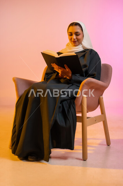 Practicing the hobby of reading, the concept of passion for reading, a portrait of a veiled Gulf Arab Omani woman wearing a colorful abaya sitting on a comfortable chair and reading a book, purposeful and entertaining books and novels, learning and education through reading purposeful books, colorful background