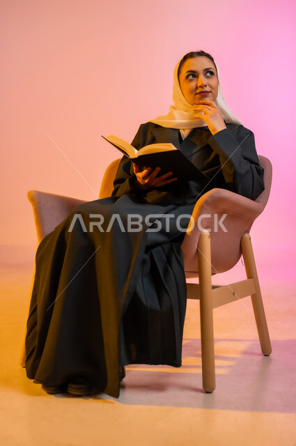 Practicing the hobby of reading, the concept of passion for reading, a portrait of a veiled Gulf Arab Omani woman wearing a colorful abaya sitting on a comfortable chair and reading a book, purposeful and entertaining books and novels, learning and education through reading purposeful books, colorful background