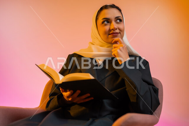 ممارسة هواية المطالعة, مفهوم الشغف بالقراءة, بورتريه لامراة عربية خليجية عمانبه محجبة ترتدي العباءة الملونة تجلس على كرسي مريح و تقوم بقراءه كتاب, كتب وروايات هادفه ومسليه ، التعلم والتثقيف من خلال قراءه الكتب الهادفه، خلفيه ملونه