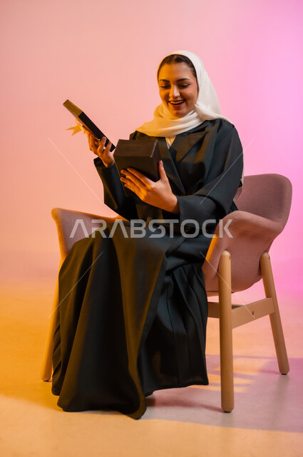 الحصول على هدية بمناسبة العيد، تقديم الهدايا في المناسبات السعيدة، بورتريه لشابة عربية خليجية عمانيه محجبة ترتدي العباءة تجلس على كرسي مريح تحمل بيدها علبة هديه مغلفة تقوم بفتحها لرؤيه مابداخلها بشغف ولهفه، الاحتفال بقدوم الأعياد والمناسبات السعيده، خلفيه ملونه