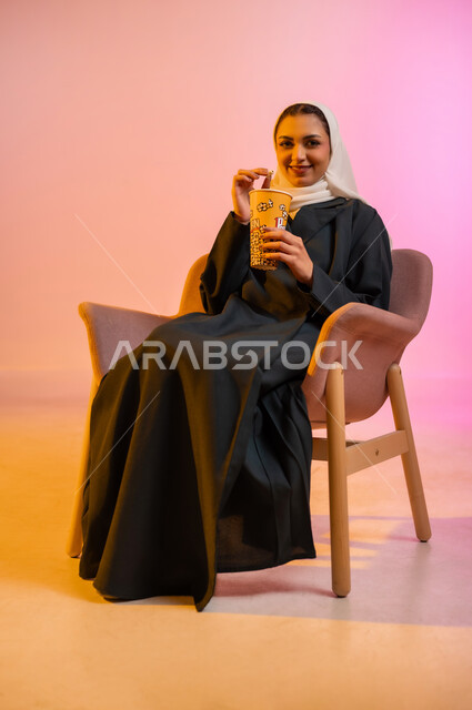 الاستمتاع بتناول السناك خلال أوقات الفراغ، بورتريه لشابه عربية عمانيه خليجية  ترتدي الحجاب والعباءة الملونه تجلس على كرسي مريح وتقوم بتناول الفشار بايماءات الدهشه والسرور،  التسلية والاستمتاع بقضاء الاوقات الممتعه بمشاهده التلفاز، خلفيه ملونه