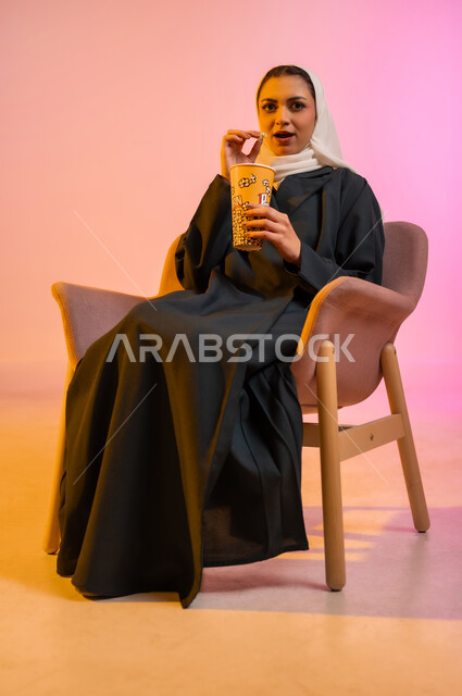 Enjoying eating snacks during leisure time, a portrait of a young Arab, Omani Gulf woman wearing the hijab and colorful abaya, sitting on a comfortable chair and eating popcorn with gestures of amazement and pleasure, having fun and enjoying spending enjoyable times watching television, colorful background.