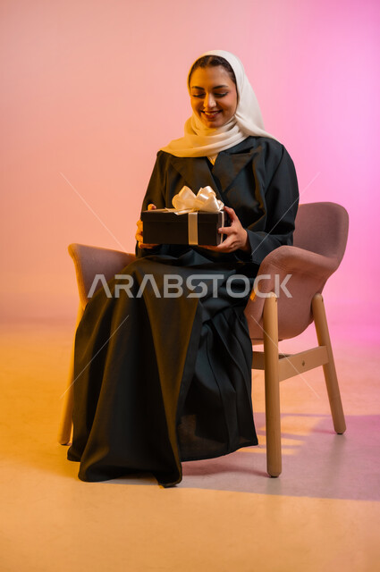 مفهوم الشكر والامتنان، تقديم الهدايا بالاعياد والمناسبات الخاصة، التعبير عن الحب والتقدير والاحترام، بورتريه لسيده عربيه خليجيه عمانيه تجلس على كرسي وتحمل بيديها صندوق هدية تنظر اليه بايماءات السعاده والسرور، خلفيه ملونه