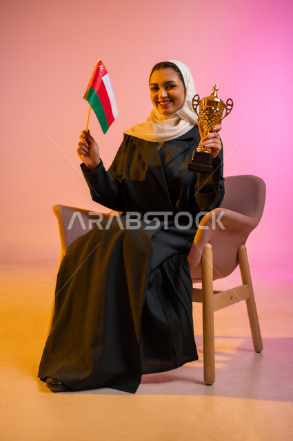 Participation in national holidays and occasions, pride in belonging to identity, celebrating the anniversary of Omani National Day, pride in Omani symbols and flag, portrait of a smiling veiled Omani Gulf Arab young woman wearing a black abaya sitting on a chair holding the flag and cup, colored background